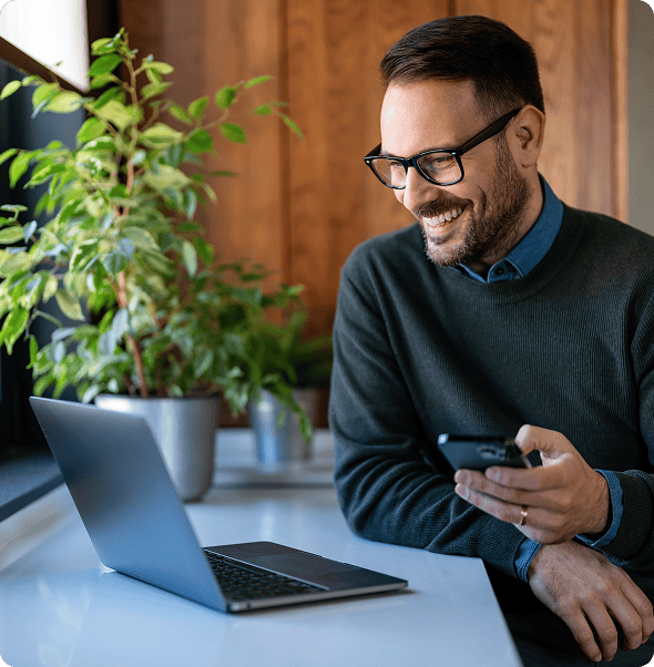 A man using a smartphone with a laptop on a desk, in a modern office setting.